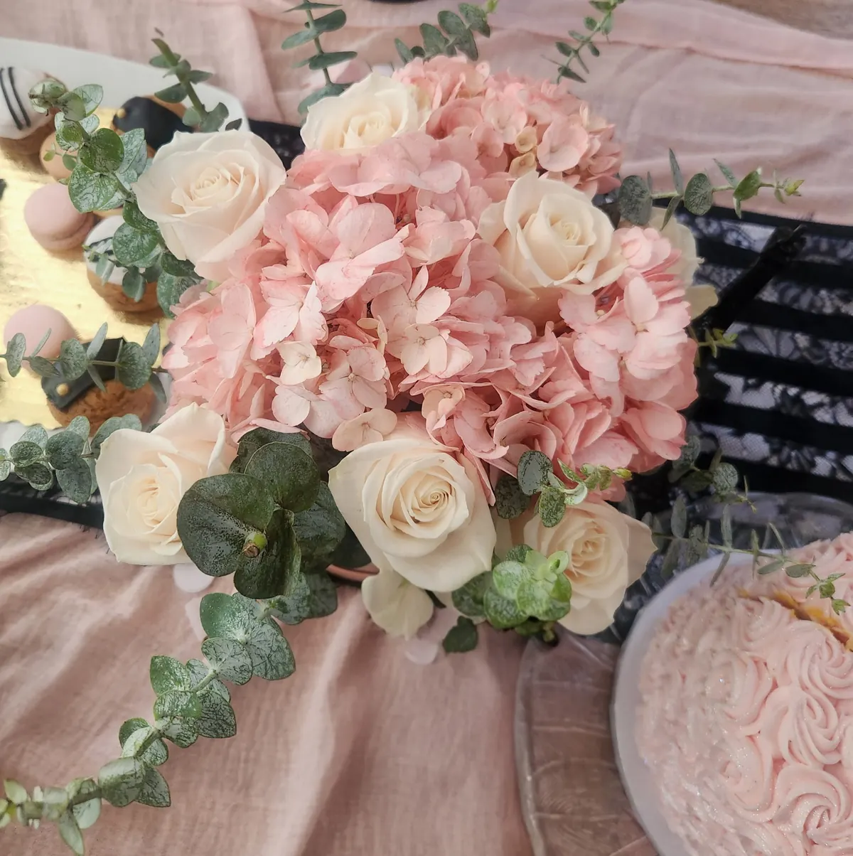 Cream roses and pink hydrangeas with trailing eucalyptus beside macarons and a pink cake