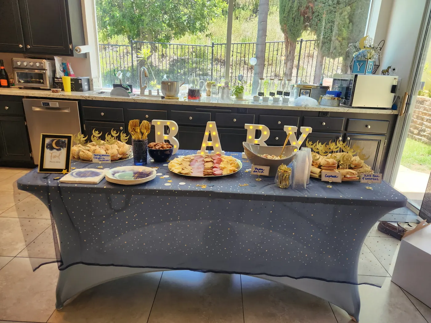 Grazing table draped in a starry blue cloth with illuminated BABY letters and labeled food trays